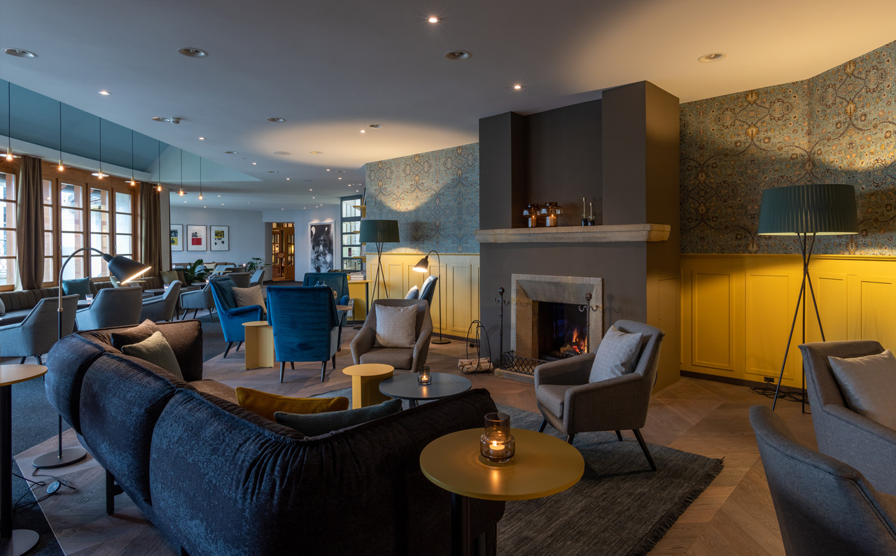 Ein grosser Lounge-Bereich mit vielen Sesseln und Sofas in Grau- und Blautönen. In der Mitte steht ein Kamin mit Feuer. Die Wände haben gelbe Holzverkleidungen und gemusterte Tapeten. Mehrere kleine Tische und Steh- sowie Tischlampen stehen im Raum. Links sind grosse Fenster mit braunen Vorhängen
