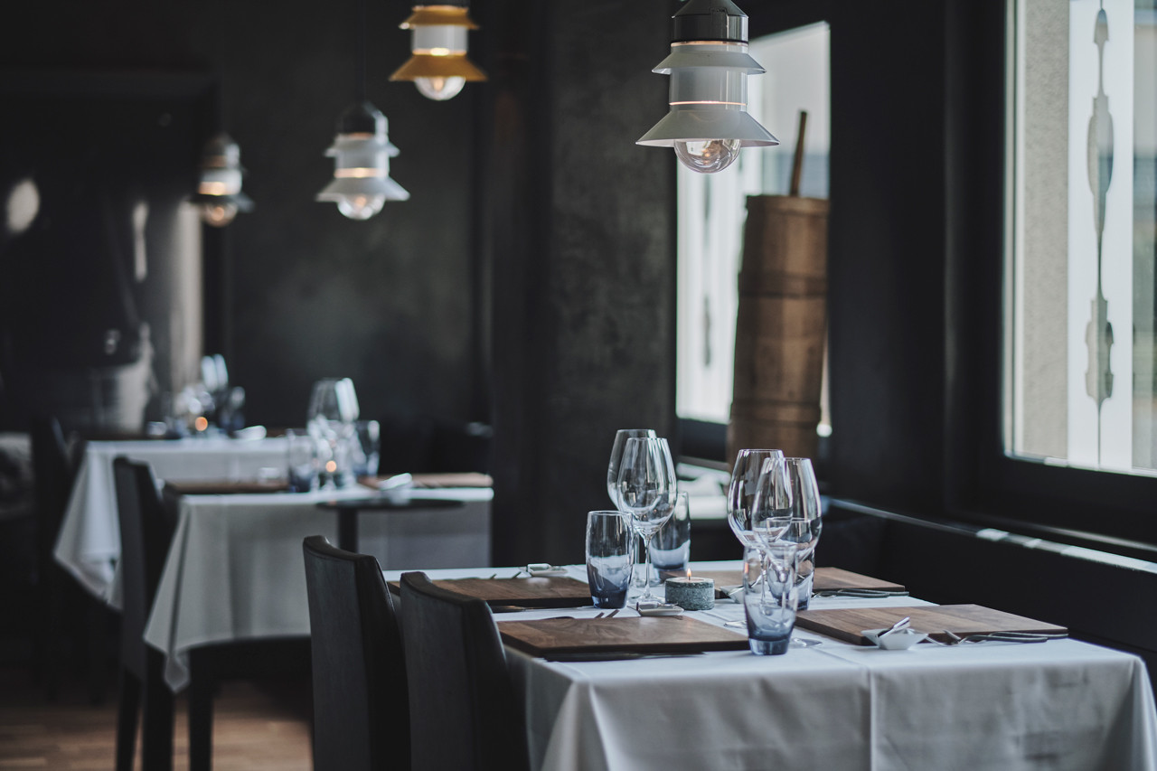 ChatGPT: Ein Restaurantbereich mit weiss gedeckten Tischen, dunklen Stühlen und mehreren Wein- und Wassergläsern auf den Tischen. Über den Tischen hängen mehrere moderne Lampen. Rechts sind grosse Fenster mit hellen Musterelementen.