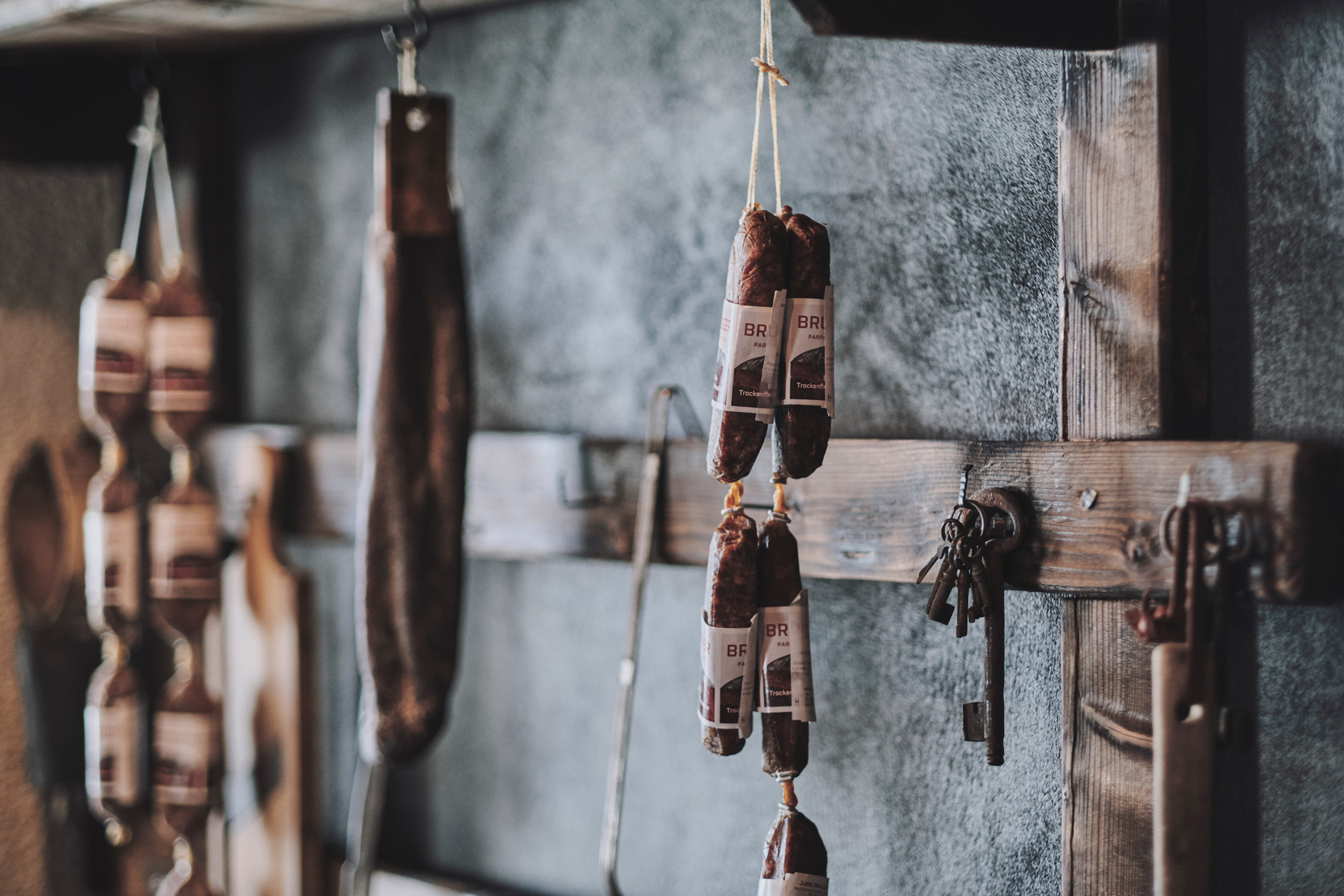 Mehrere getrocknete Würste hängen an Haken vor einer dunklen Wand. Daneben befinden sich alte Holzleisten und daran befestigte alte Metallschlüssel.