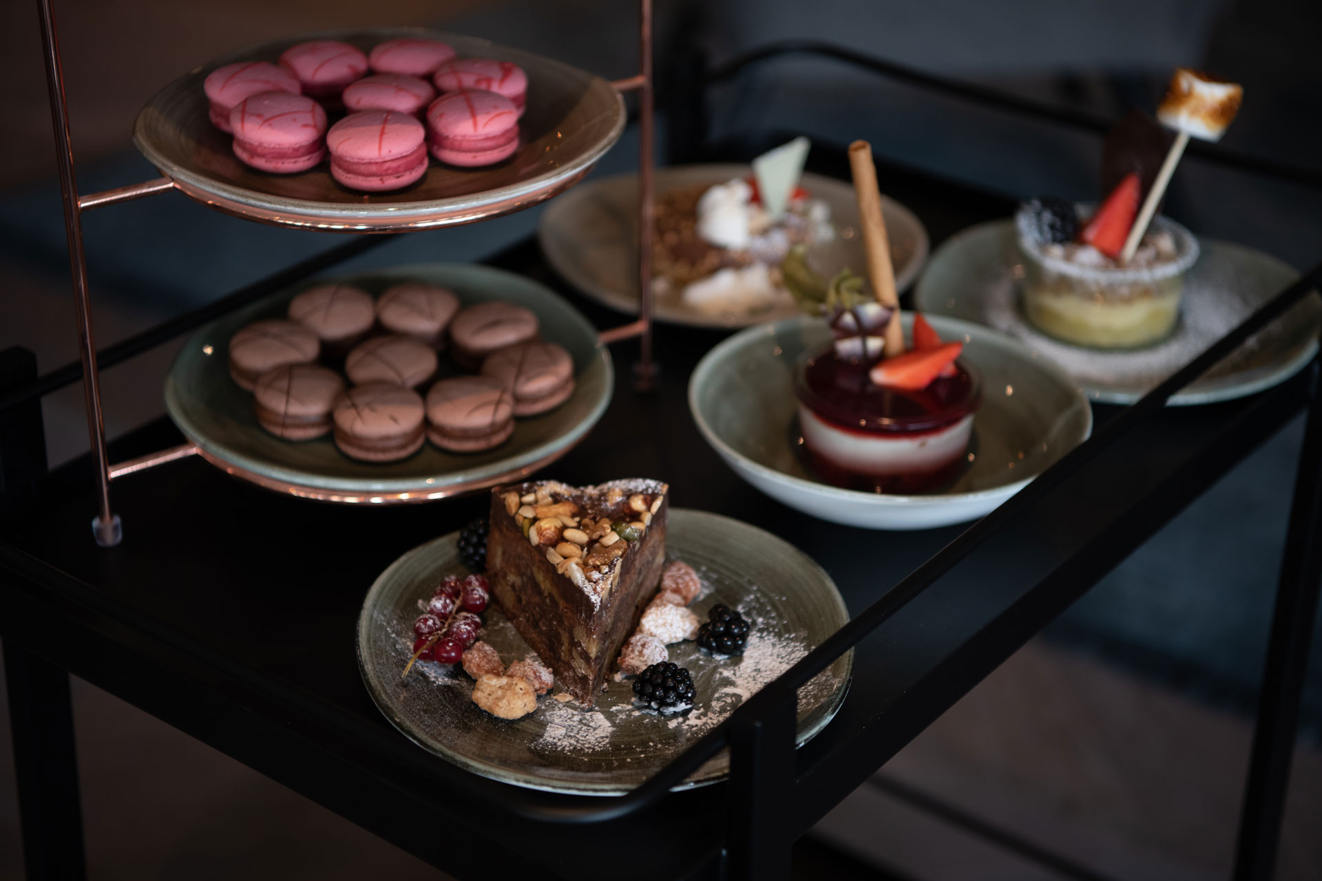 Auswahl an Desserts auf einem Servierwagen: Macarons, Tortenstück mit Nüssen, Beeren und verschiedene kleine Kuchen auf Tellern.