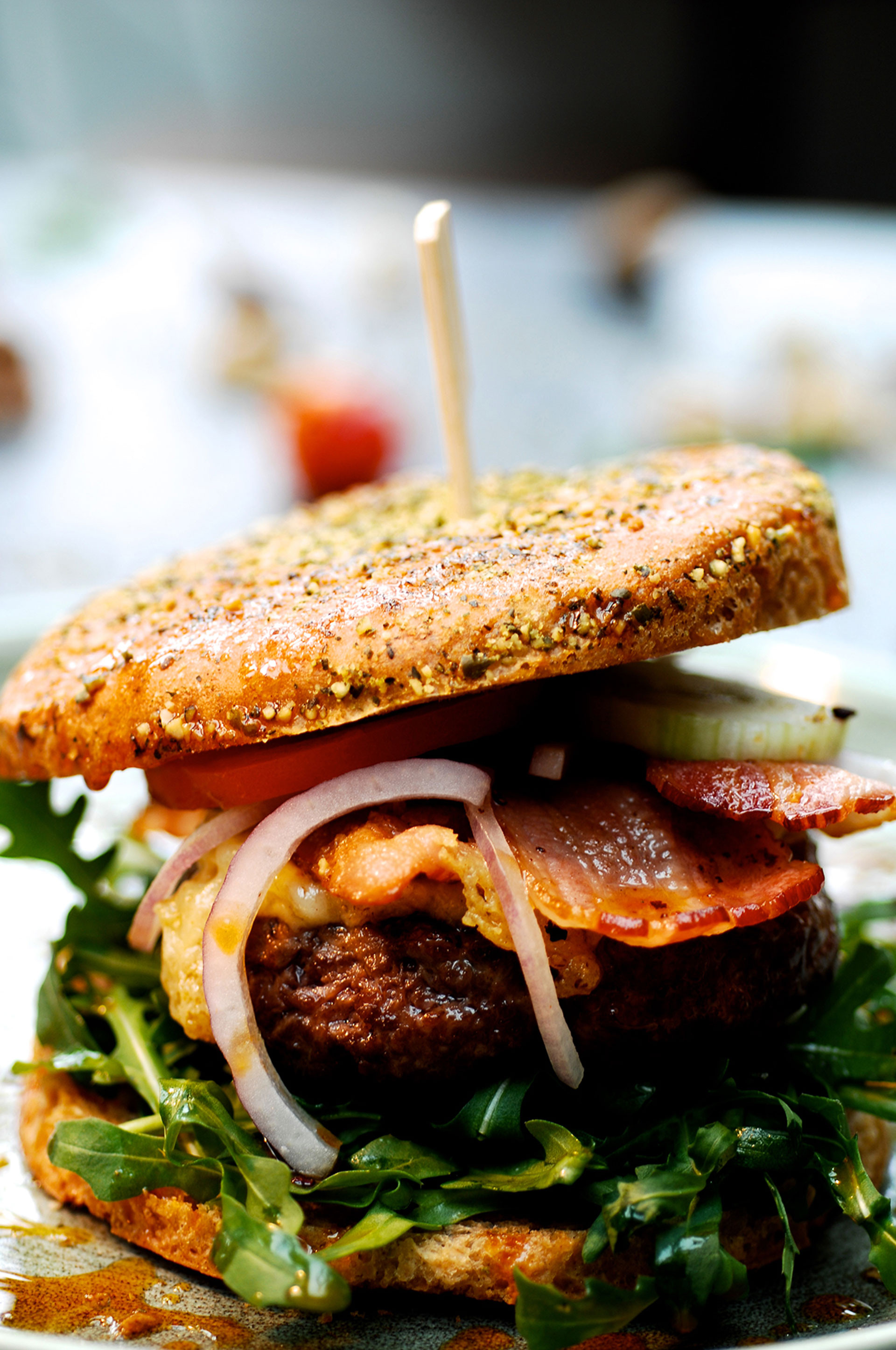 Ein Burger mit Brötchenhälften, belegt mit Rucola, einem Fleischpatty, Käse, gebratenem Speck, Tomatenscheiben, roten Zwiebeln und Gurkenscheiben. Ein Holzstäbchen steckt durch die Mitte.