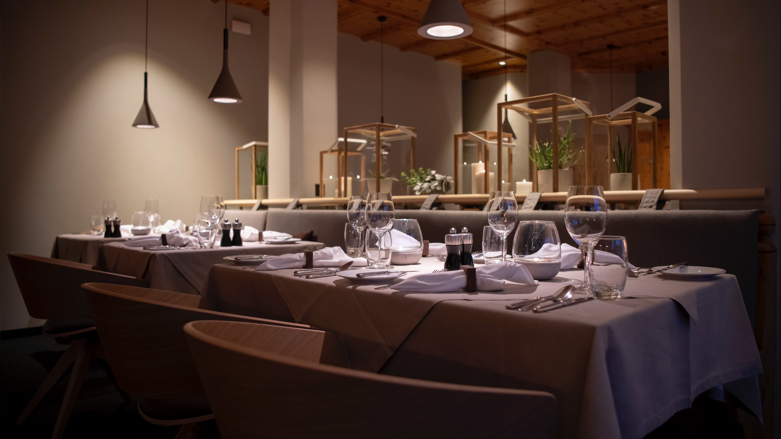 Mehrere gedeckte Restauranttische mit weißen Stoffservietten, Gläsern, Besteck und Salz-/Pfeffermühlen. Oben hängen schmale Pendelleuchten. Im Hintergrund stehen Holzrahmen mit Pflanzen und Kerzen auf einer langen Rückenlehne.