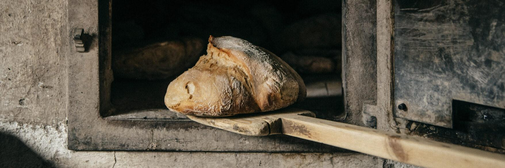 ChatGPT: Ein Laib Brot liegt auf einem hölzernen Brotschieber und wird gerade aus einem alten Stein- oder Holzofen gezogen. Der Ofen wirkt dunkel und rustikal, mit sichtbaren Gebrauchsspuren.
