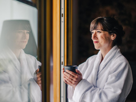 Frau im Bademantel mit einer Tasse Tee blickt entspannt aus dem Fenster im Wellnessbereich des Hotel Schweizerhof Lenzerheide