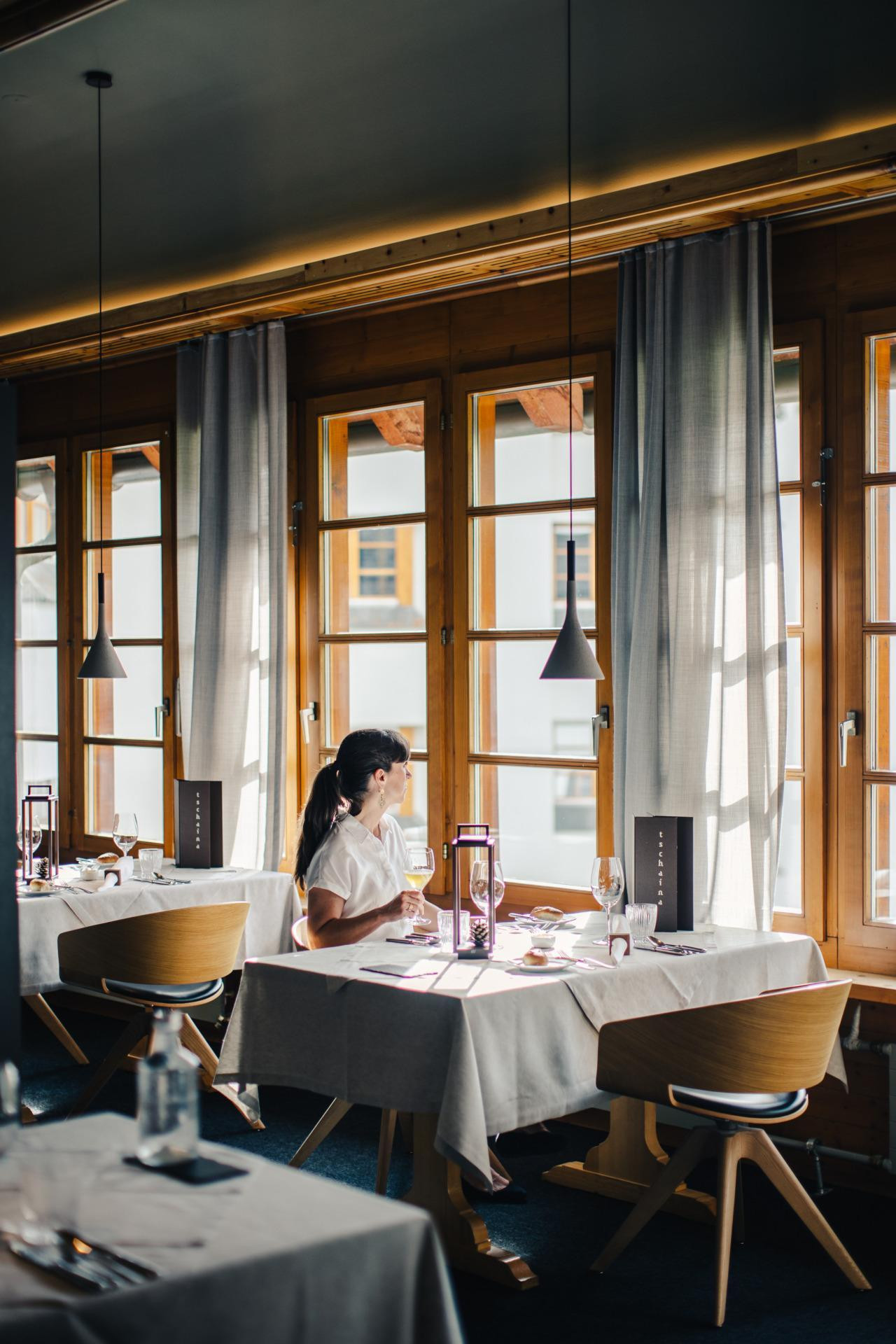 Frau genießt entspannten Moment am Fenster im Restaurant im Hotel Schweizerhof Lenzerheide bei Tageslicht und Alpenblic