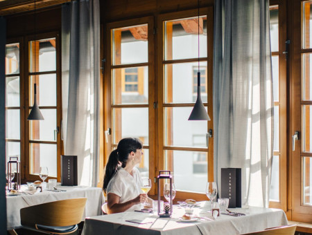 Frau genießt entspannten Moment am Fenster im Restaurant im Hotel Schweizerhof Lenzerheide bei Tageslicht und Alpenblic