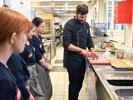Koch zeigt Schülerinnen des Behind the Scenes Projekts im Hotel Schweizerhof Lenzerheide das fachgerechte Schneiden von Zutaten