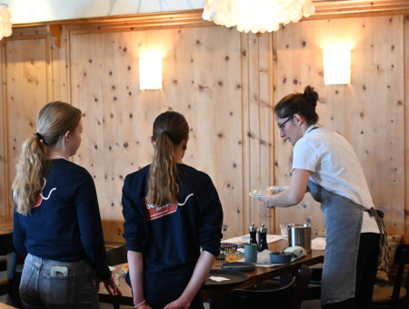 Service-Mitarbeiterin erklärt zwei Schülerinnen des Behind the Scenes Programms den Ablauf beim Servieren im Hotel Schweizerhof Lenzerheide