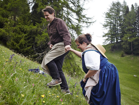 Frau in Tracht hilft Koch auf blühender Alpiese – gelebte Regionalität und Gemeinschaft im Scalottas Terroir Lenzerheide