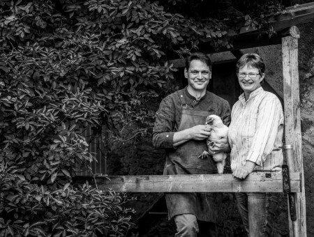 „Schwarzweißaufnahme zweier Menschen mit Huhn vor Bauernhaus – partnerschaftliche Zusammenarbeit im Scalottas Terroir Lenzerheide