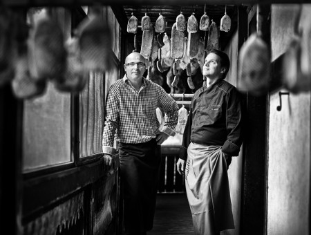 Schwarzweißaufnahme zweier Männer zwischen hängenden Trockenfleisch-Spezialitäten im Scalottas Terroir Lenzerheide – handwerkliche Tradition und Regionalität