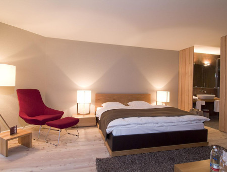 Geräumiges Zimmer Alpenchic im Hotel Schweizerhof Lenzerheide mit Doppelbett, rotem Loungesessel und warmem Licht für Wohlfühlatmosphäre