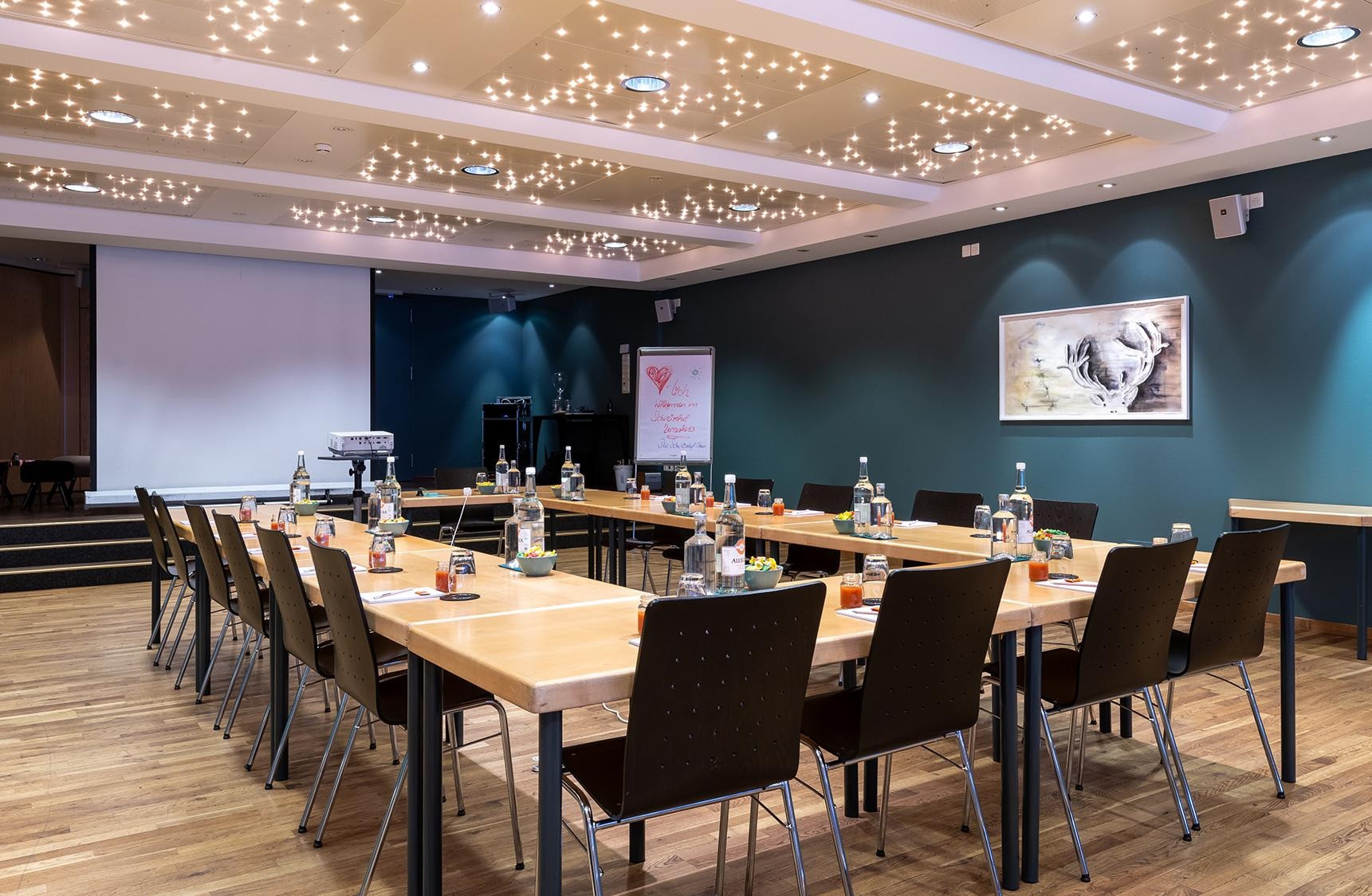 Seminarraum mit Tischen in der Mitte mit Wasserflaschen und Schreibblöcke sowie Stifte. Vorne im Raum ein Beamer mit einer Weissen Leinwand.