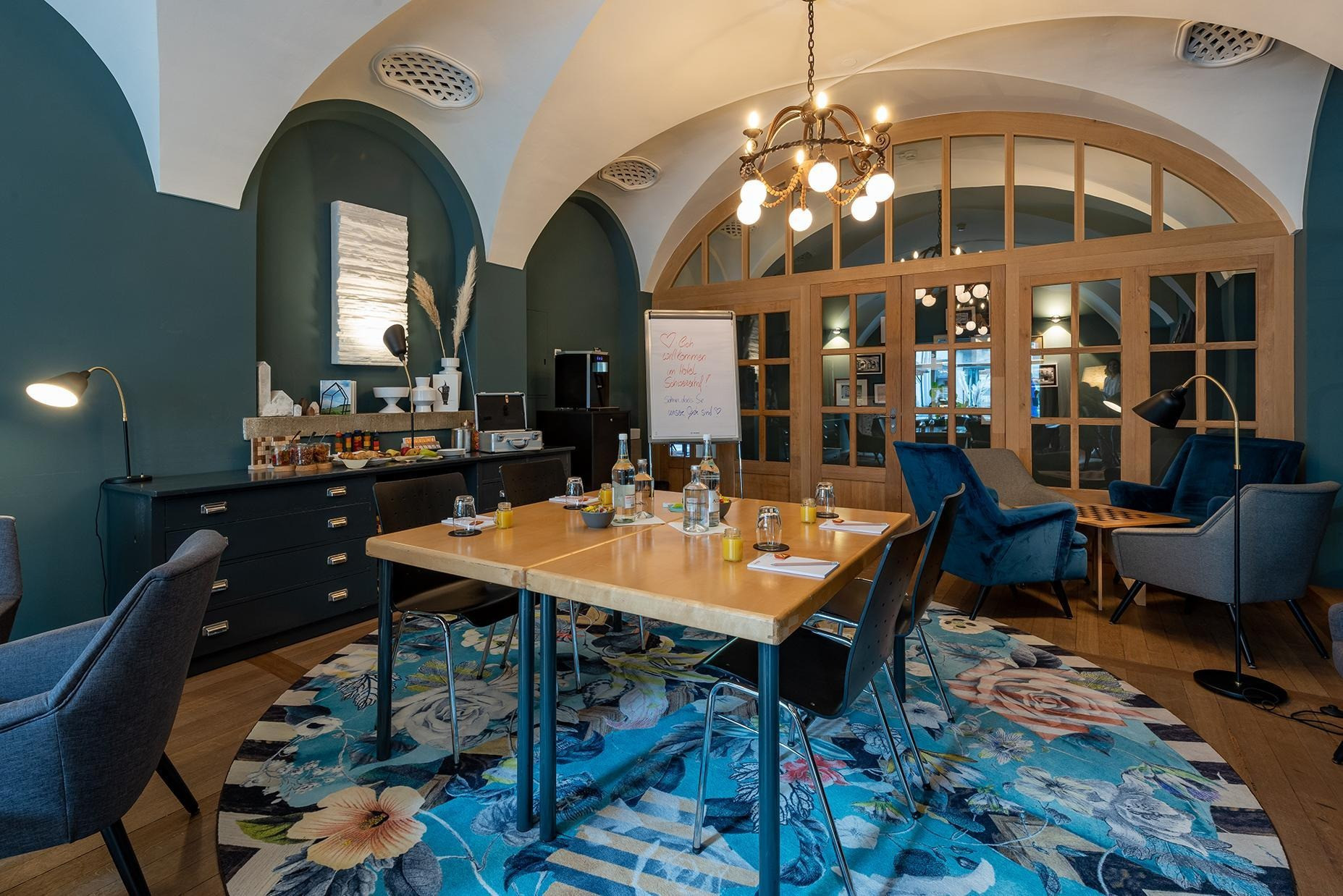 Raum mit einem Kronleuchter und zwei Tische in der Mitte mit Stiften und Schreibblöcken. Im Hintergrund eine Sitzlounge mit einem Tisch mit einem Schachbrett und eine Ablage mit Obst und Trockenfrüchten und Nüssen.