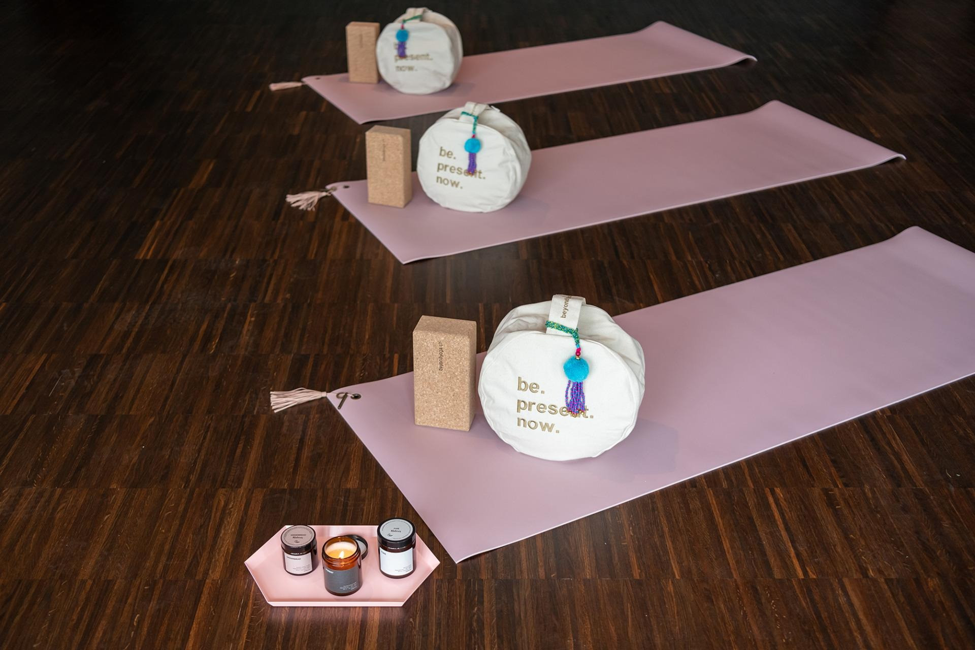 Yogamatten-Setup im Fitnessbereich des Hotelsmit rosa Matten, Meditationskissen „be. present. now.“, Korkblöcken und Kerzen auf dunklem Holzboden.