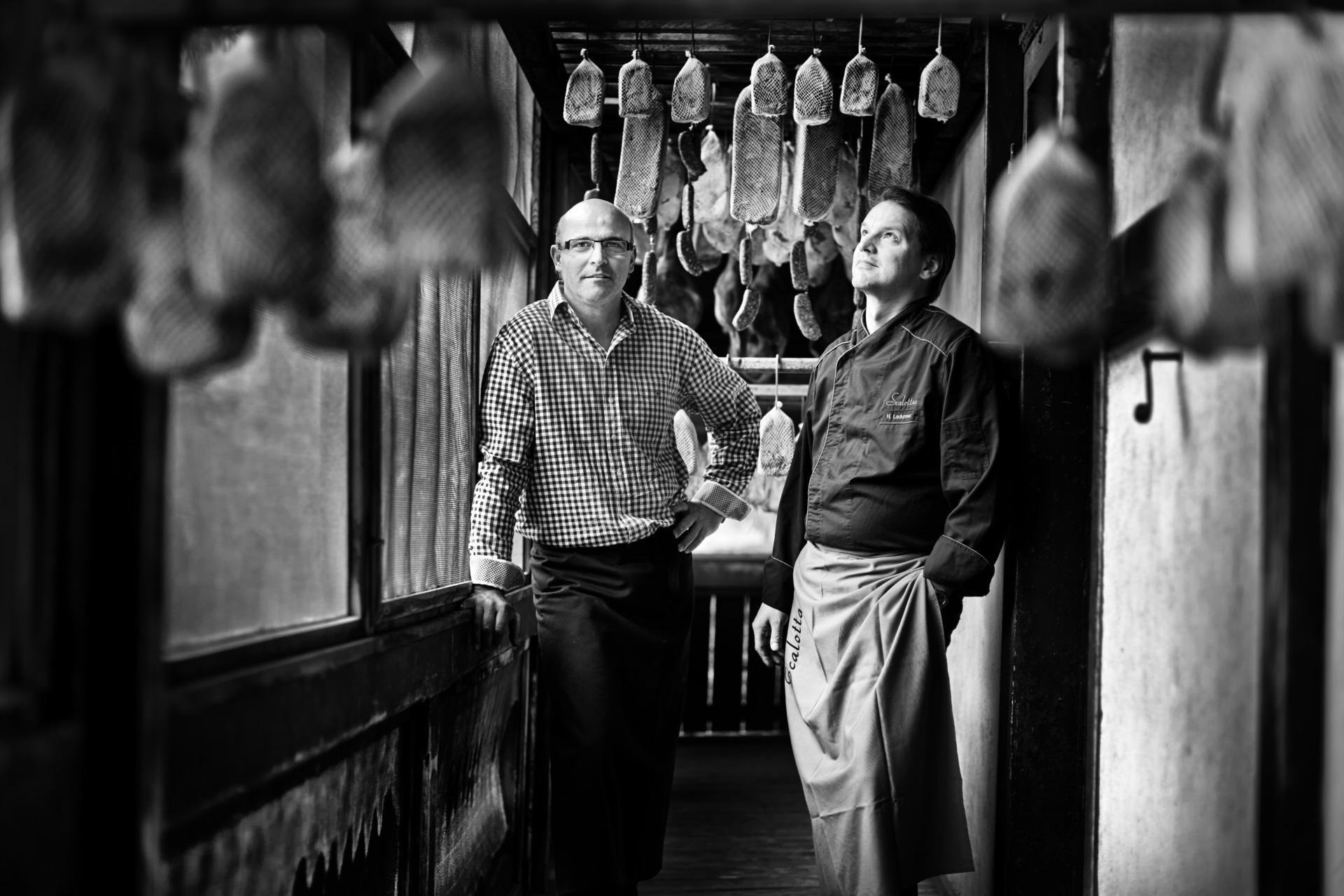 Zwei Mitarbeiter stehen in einer traditionellen Räucherkammer im Scalottas Terroir in Lenzerheide zwischen hängenden Fleisch- und Wurstspezialitäten.