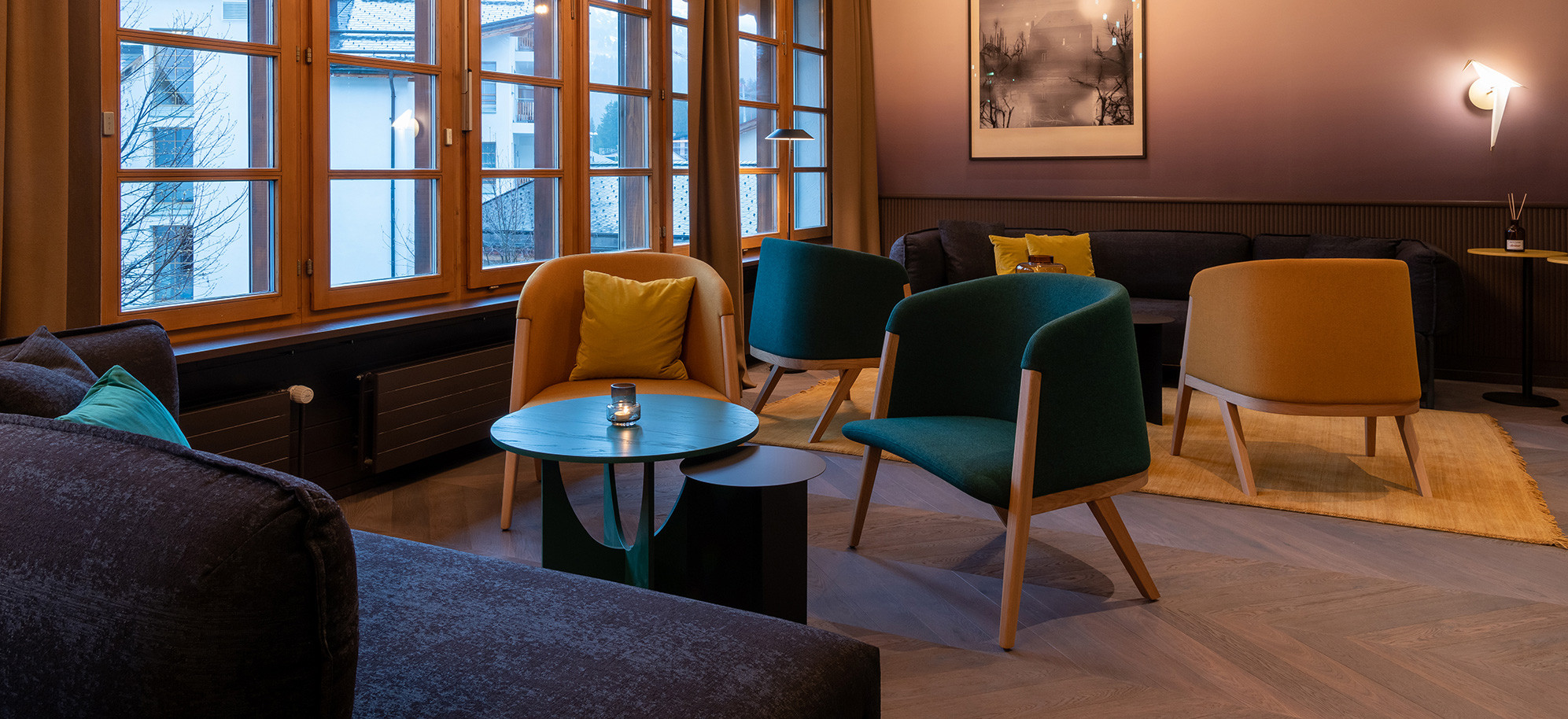 Gemütlicher Lounge-Bereich mit farbigen Sesseln, Sofas, kleinen Tischen und grossen Holzfenstern in einem modernen Innenraum.