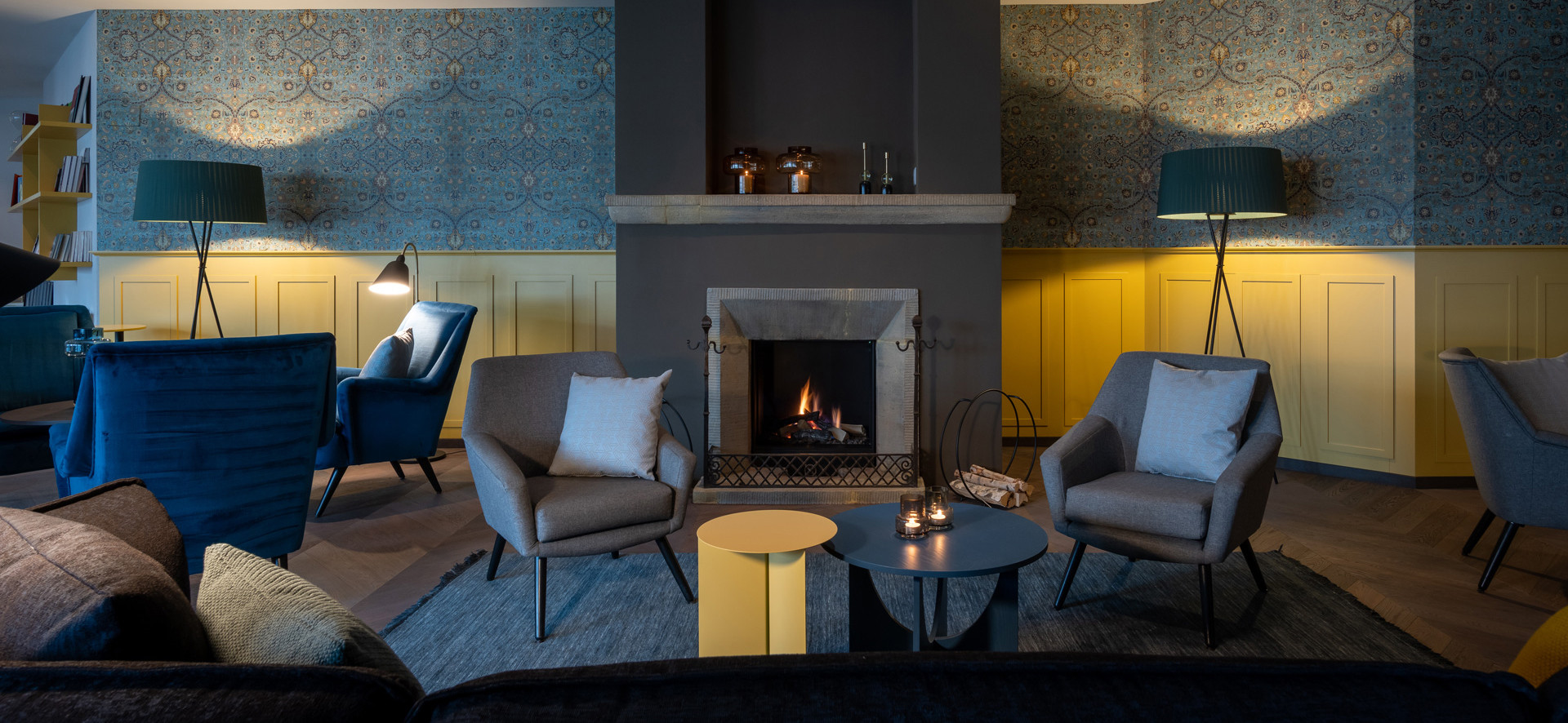 Moderner Loungebereich mit Kamin, grauen und blauen Sesseln, gelben Wandpaneelen, gemusterter blauer Tapete und kleinen Beistelltischen.