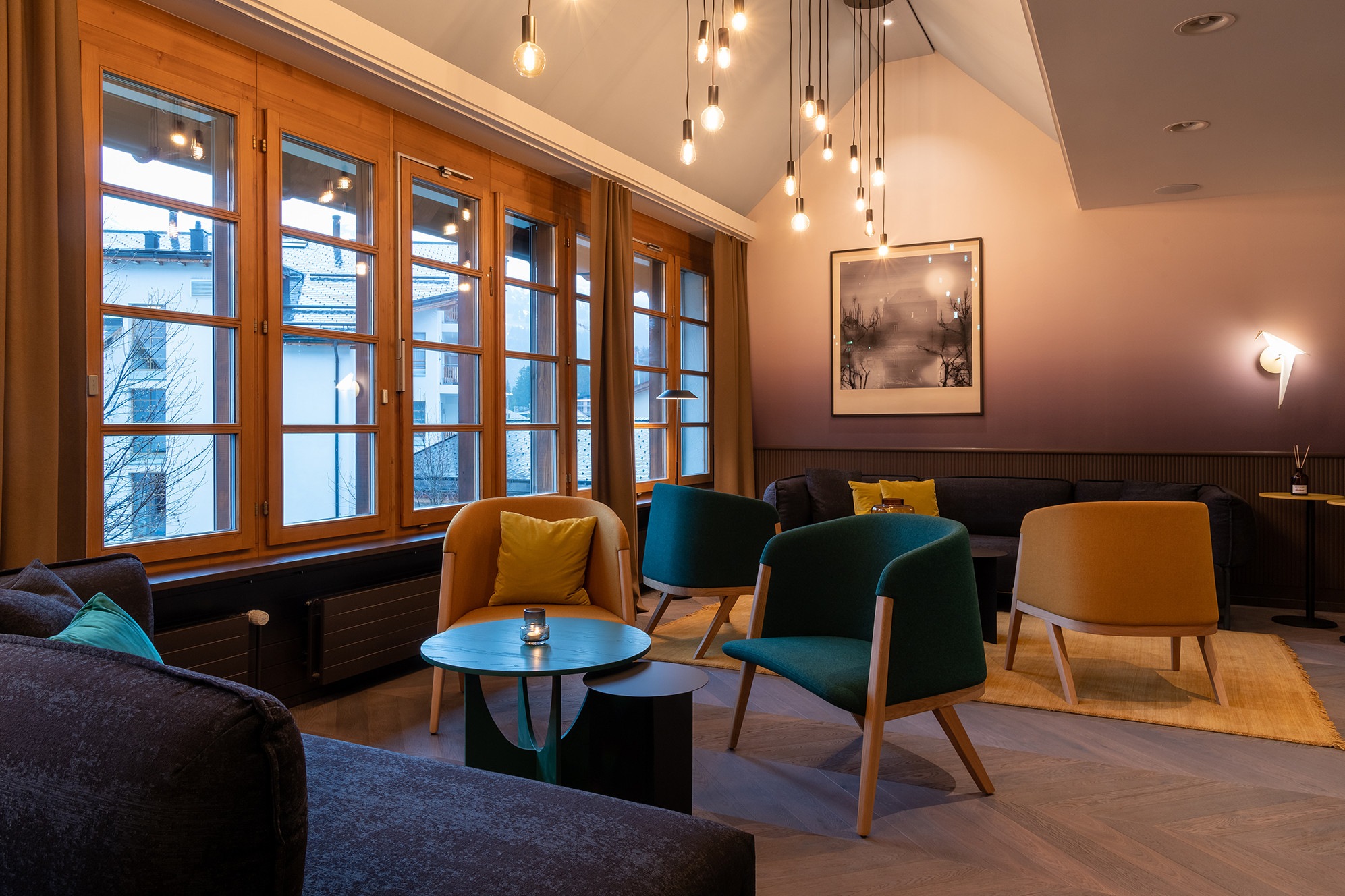 Moderner Loungebereich mit grossen Holzfenstern, farbigen Designerstühlen, kleinen Beistelltischen, Sofas und dekorativen Hängeleuchten.