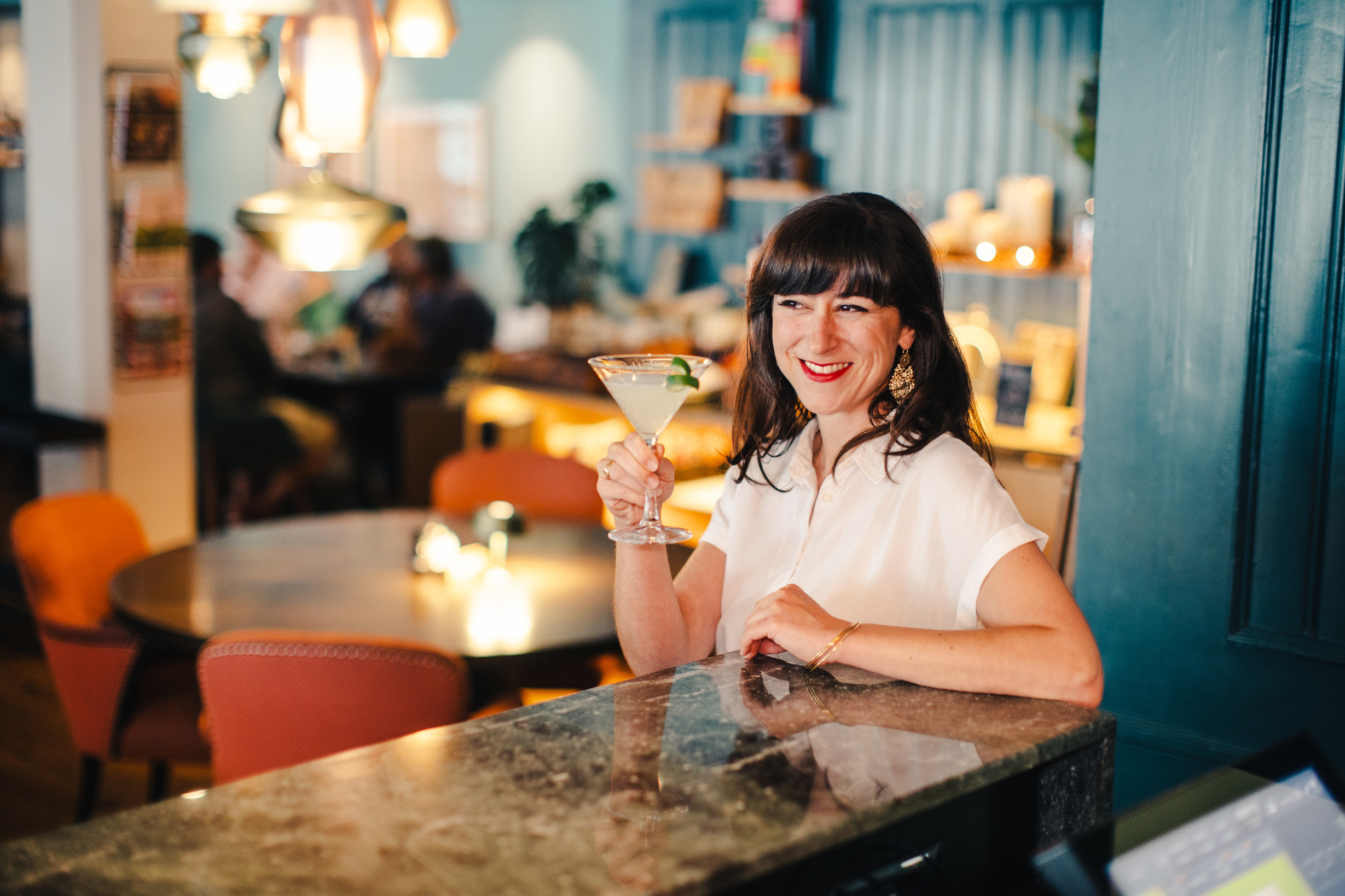 Frau an der Hotelbar mit einem Cocktailglas, lächelnd und umgeben von warmem Lounge-Ambiente mit stilvoller Beleuchtung.