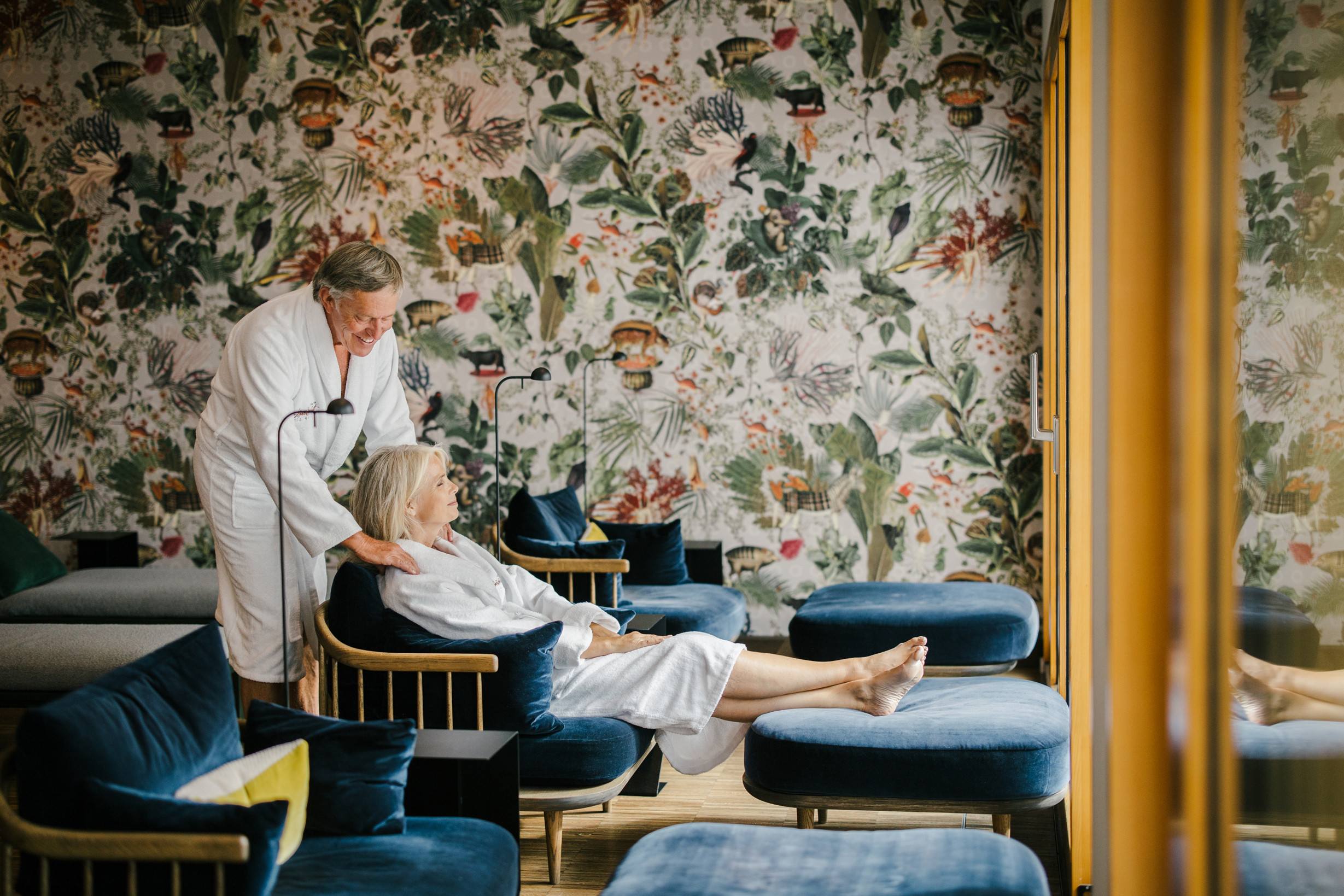 Pärchen geniesst in Spabereich die Ruhelounge. Die Frau liegt auf einer Liegefläche während der Mann fröhlich ihre Schultern massiert. Im Hintergrund eine Florate Vintage Fassade.