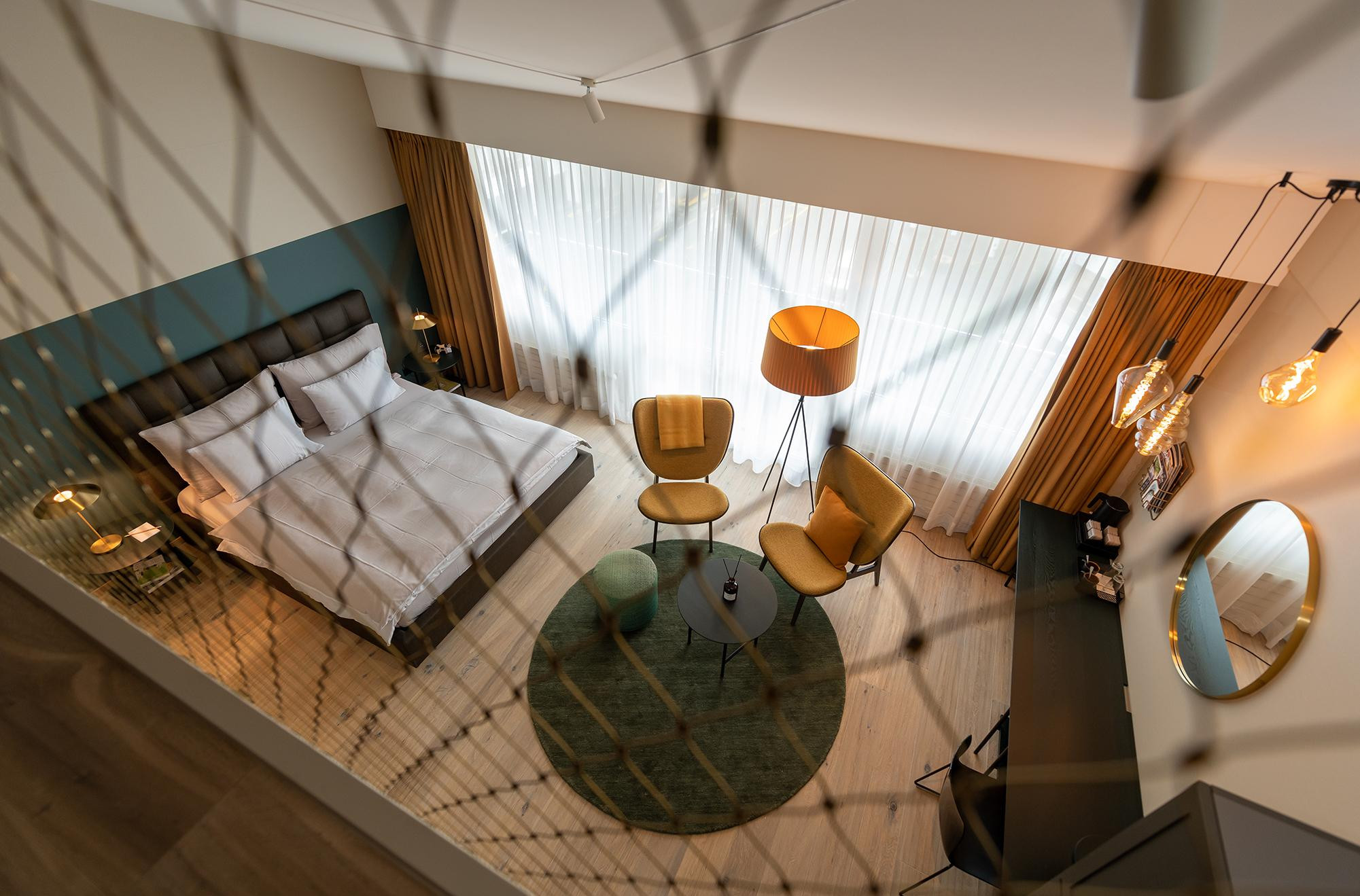 Modernes, helles Hotelzimmer mit großem Doppelbett, gemütlicher Sitzecke in warmen Gelb- und Grüntönen, bodentiefen Fenstern und stylischer Beleuchtung – aufgenommen von der Galerieebene.