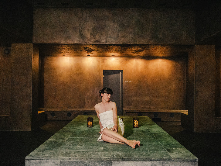 Frau sitzt im Hamambereich auf einer Liege- und Sitzfläche mit einem Handtuch. Um sie herum Kerzenlichter.