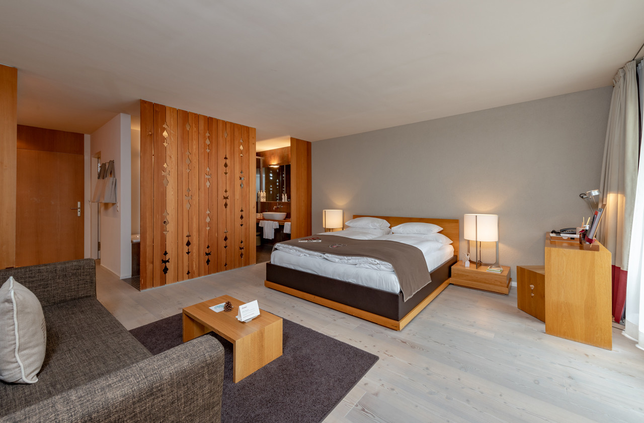 Grosszügiges Doppelzimmer im Hotel Schweizerhof Lenzerheide mit modernem Holzdécor, gemütlichem Kingsize-Bett, offener Badlösung mit Holztrennelement und komfortabler Sitzecke für entspannte Aufenthalte.