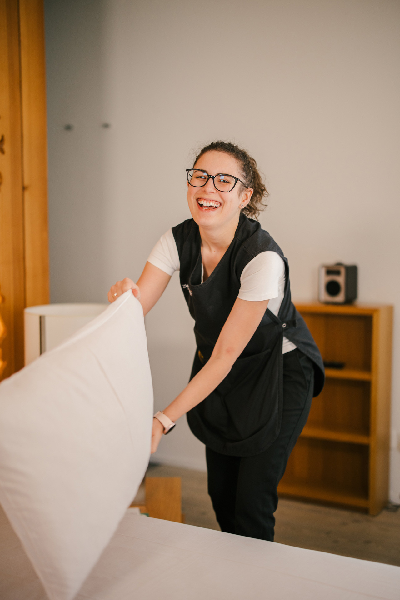 Mitarbeiterin beim Bettenmachen im Hotelzimmer, lächelnd und mit einem Kissen in der Hand.