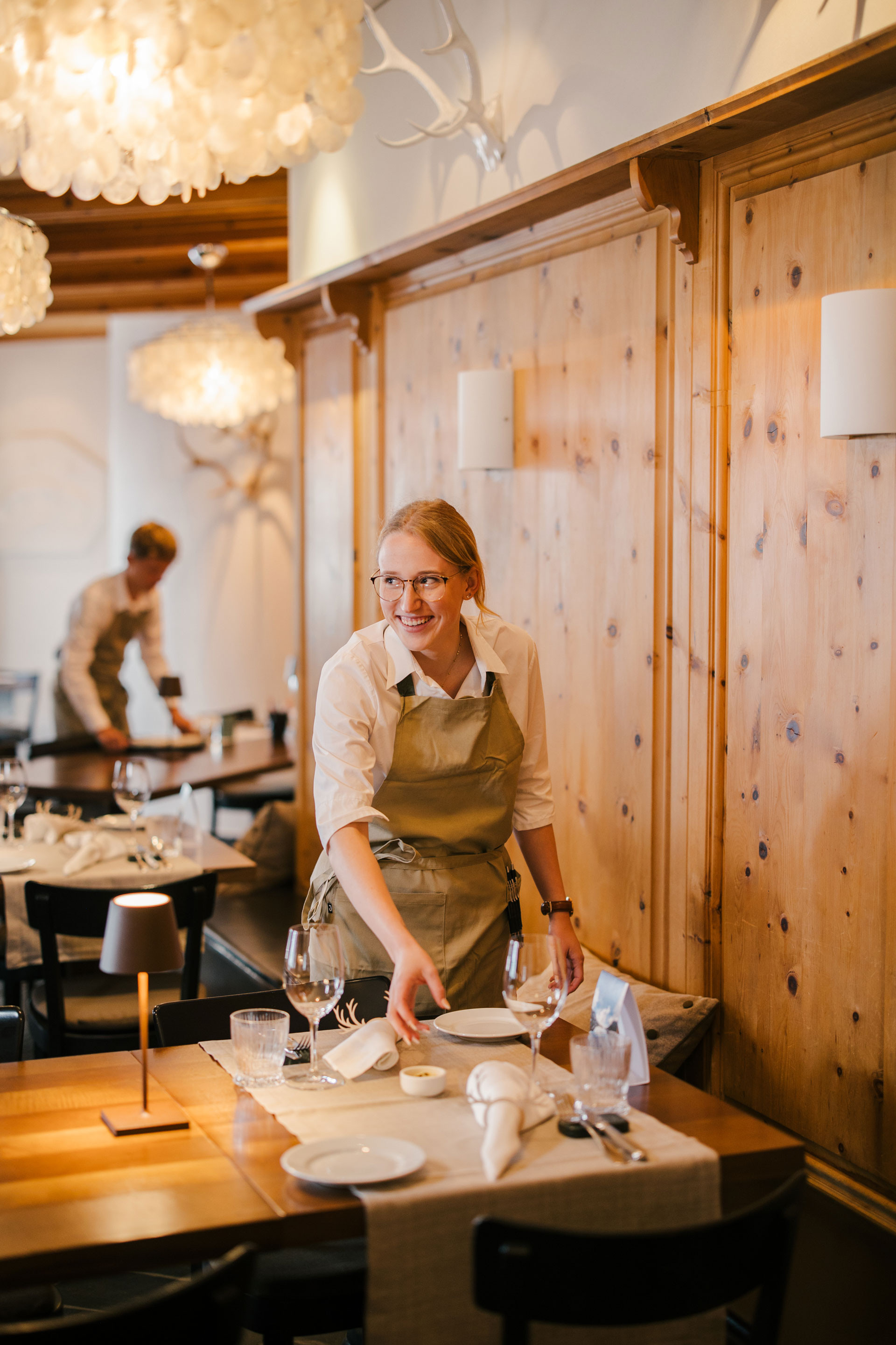 Servicemitarbeiterin richtet einen gedeckten Restauranttisch und lächelt, während im Hintergrund eine weitere Person Tische vorbereitet.
