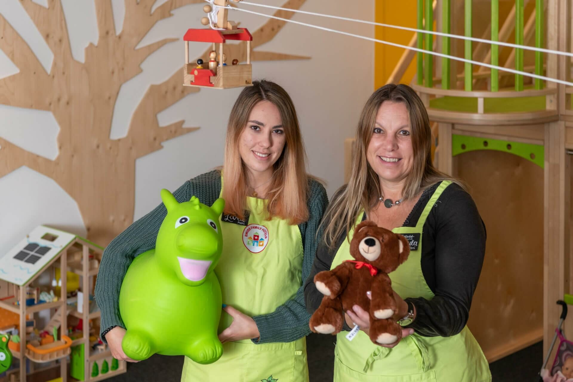 ei Mitarbeiterinnen der Kinderbetreuung in grünen Schürzen halten ein grünes Hüpfspielzeug und einen Teddybären, umgeben von liebevoll gestalteten Spielbereichen.