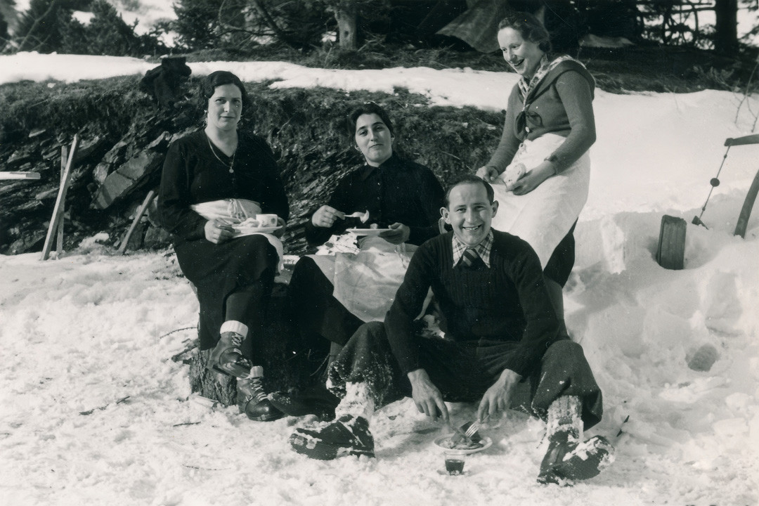 Historische Schwarz-Weiss-Aufnahme einer fröhlichen Gruppe von vier Personen, die im verschneiten Gelände gemeinsam eine Pause mit warmen Getränken und Essen geniessen.