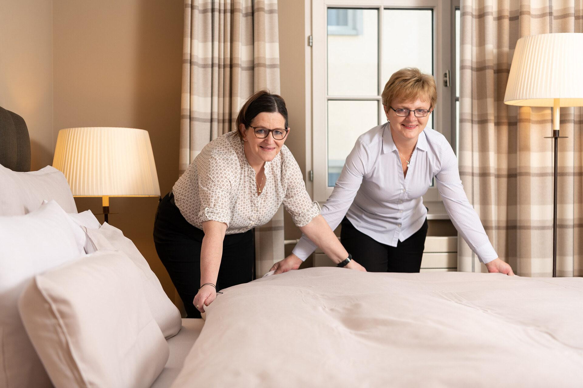Zwei Mitarbeiterinnen des Hotel Schweizerhof Lenzerheide richten gemeinsam ein frisch bezogenes Bett in einem hellen, gemütlichen Gästezimmer her.