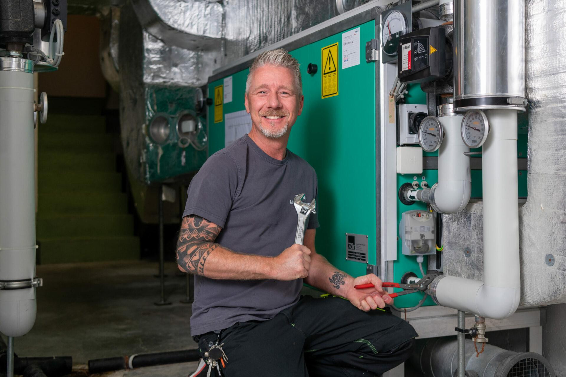 Ein Haustechniker des Hotel Schweizerhof Lenzerheide arbeitet lächelnd an einer großen grünen Heizungs- und Lüftungsanlage.