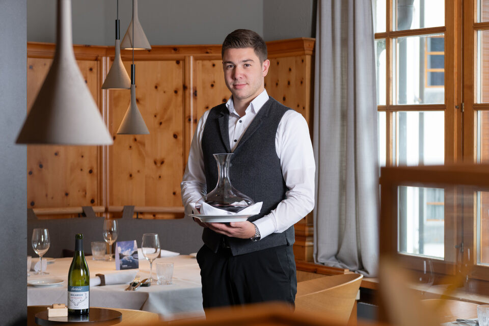 Service-Mitarbeiter im Restaurant des Hotel Schweizerhof Lenzerheide präsentiert eine stilvolle Weindekantierkaraffe – professioneller Service und hochwertige Gastronomie in alpinem Ambiente.