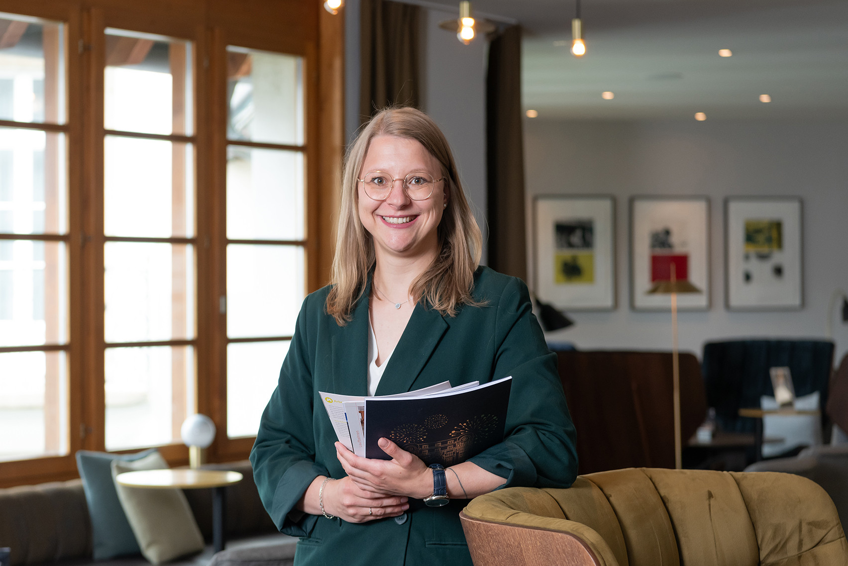 Mitarbeiterin des Hotel Schweizerhof Lenzerheide steht lächelnd in der stilvollen Lobby und hält Unterlagen in den Händen – professionelles Porträt für Gastfreundschaft und Servicequalität.