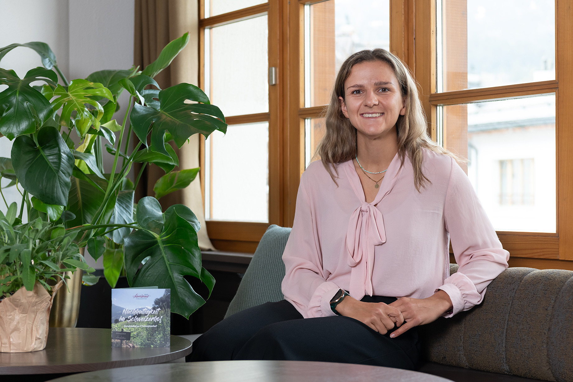 Mitarbeiterin des Hotel Schweizerhof Lenzerheide sitzt lächelnd in der Lobby vor grossen Holzfenstern, umgeben von grünen Pflanzen – Porträtaufnahme für Nachhaltigkeit und Gastfreundschaft.