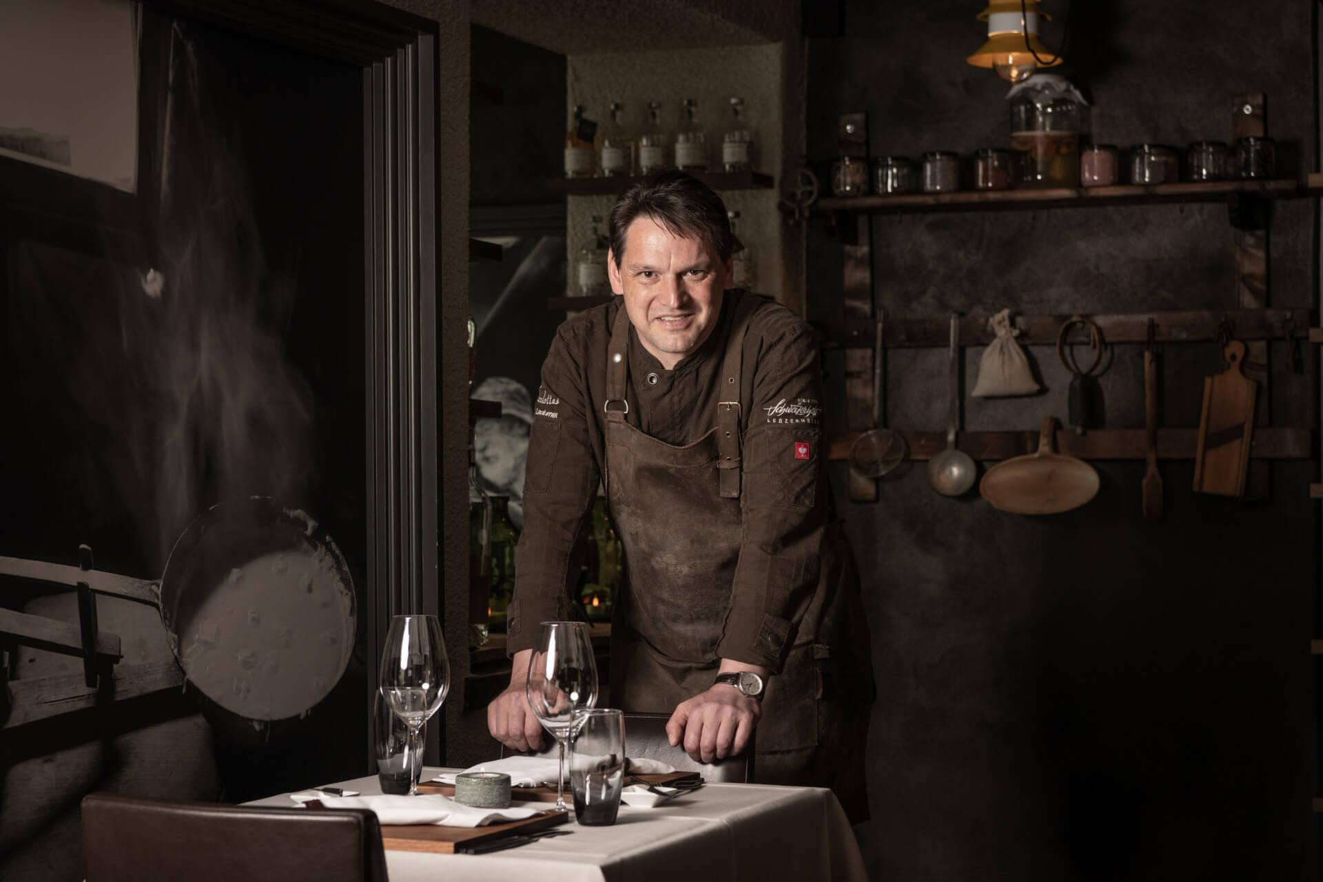 Ein Koch des Scalottas Terrior der sich mit den Händen am Stuhl abstützt. Vor ihm ein bedeckter Tisch mit Weingläser einer Kerze und Besteck.