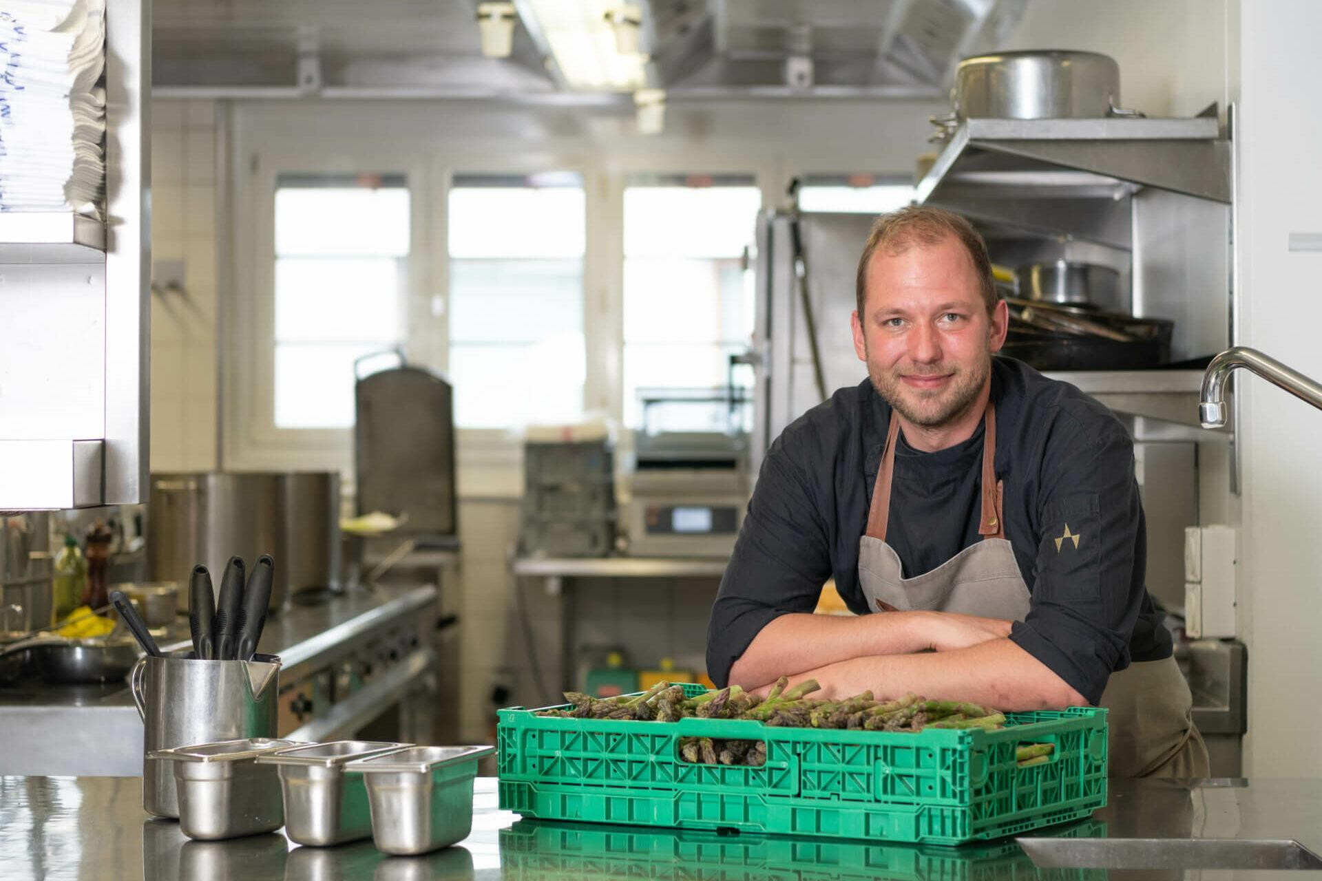 Bild eines Mitarbeiter beziehungsweise des Koches der in der Küche steht und vor sich eine Box mir frischen Spargelb hat.