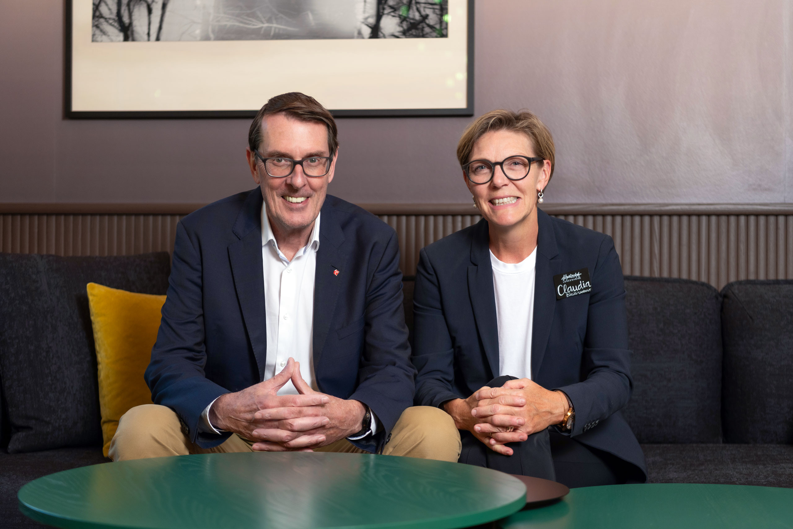 Frau und Herr Züllig lachen in die Kamera und sitzen auf einer Sitzbank im inneren des Hotels