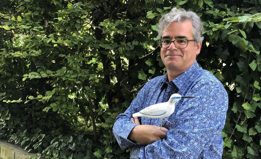 Mann der vor einem Gebüsch steht und ein Holzvogel in der Hand hält.