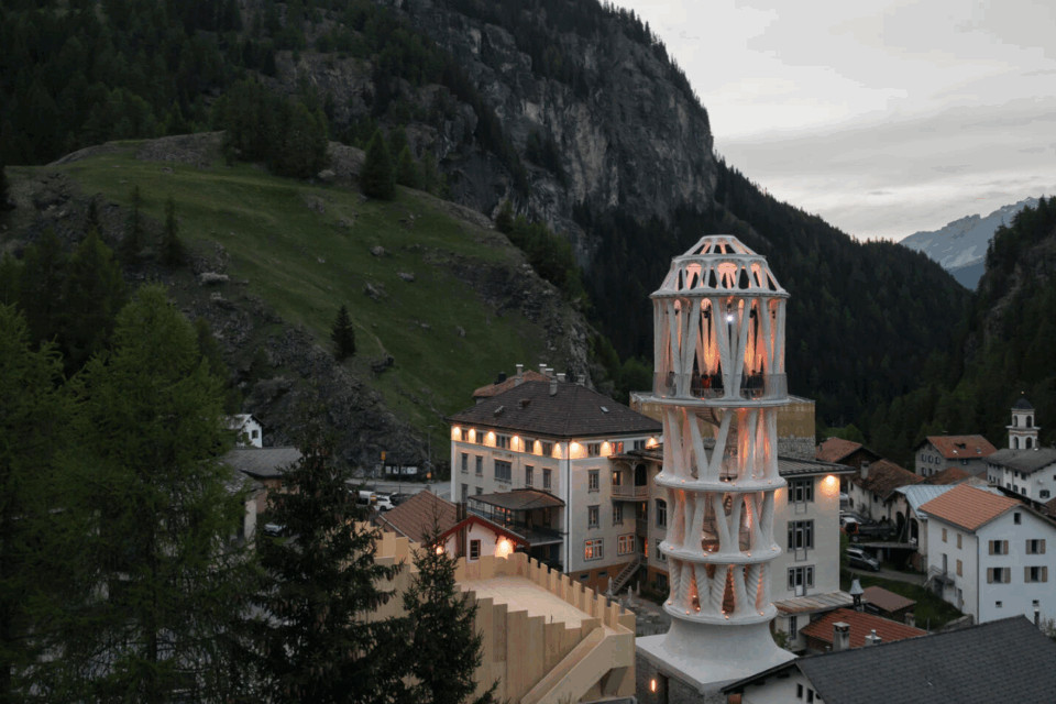 Beleuchteter, futuristischer Turm in Mulegns vor steilen Berghängen und traditionellen Häusern in der Abenddämmerung.