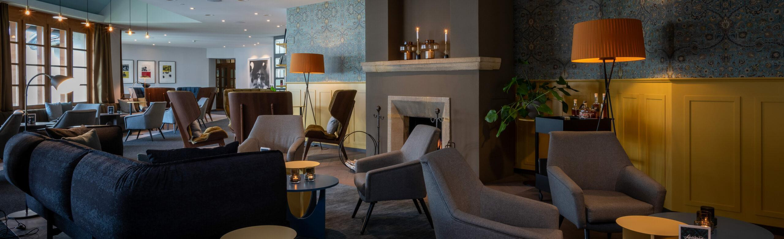 Elegante Hotel-Lounge im Schweizerhof Lenzerheide mit modernen Sesseln, Sofas, Kamin, warmem Ambientelicht, dekorativer Tapete und gemütlicher Atmosphäre für entspannte Aufenthalte.