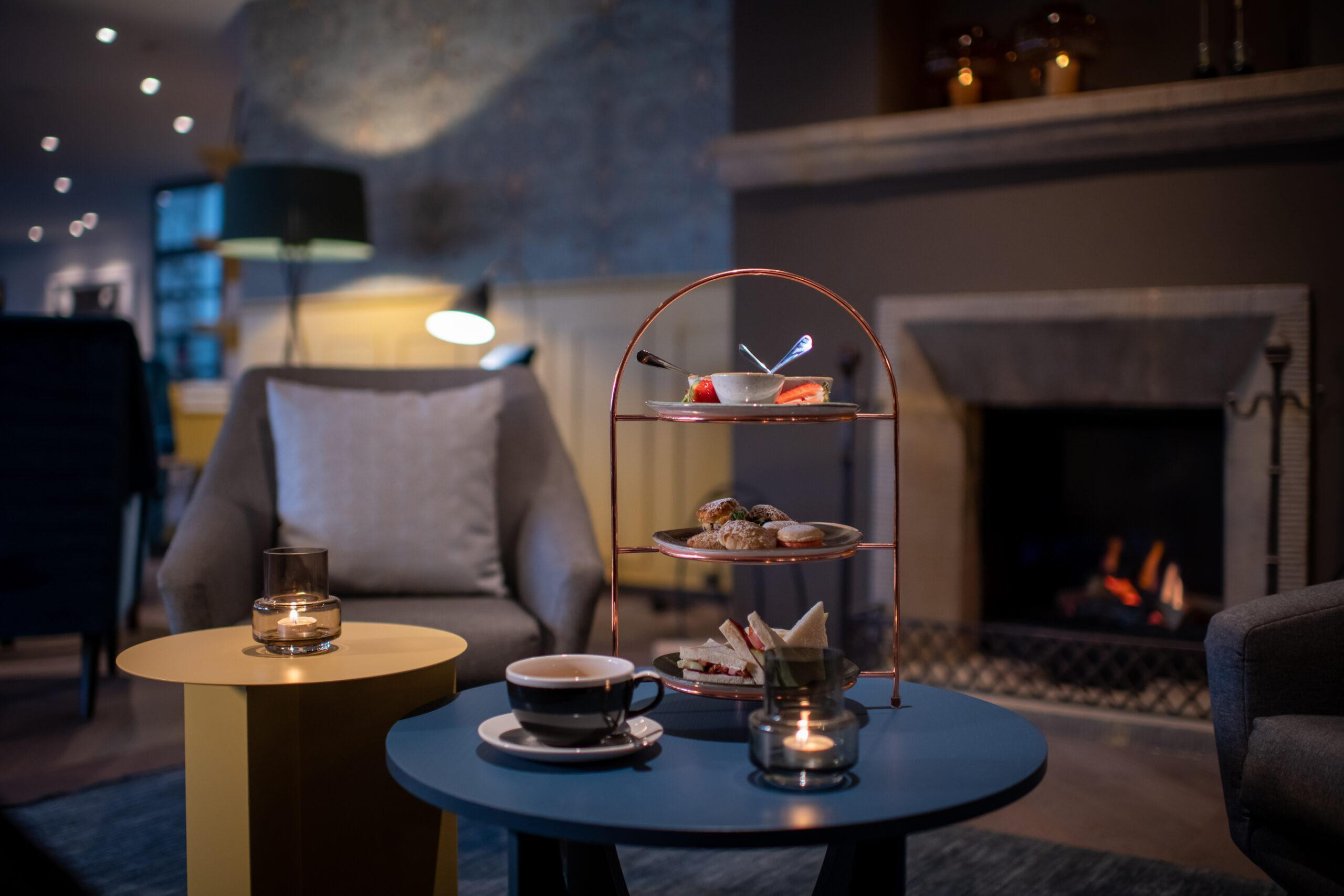 Gemütliche Lounge im Schweizerhof Lenzerheide mit Afternoon-Tea-Etagere, Sandwiches, Gebäck, Teetasse und Kerzenlicht vor knisterndem Kaminfeuer für entspannte Genussmomente.
