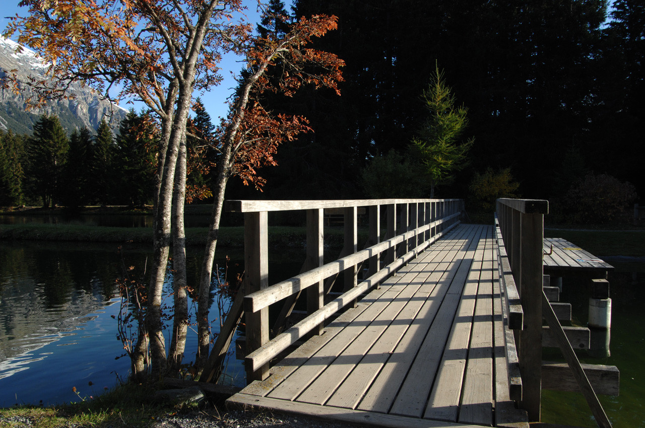 Holzsteg über einem ruhigen See, flankiert von herbstlich gefärbten Bäumen und umgeben von Berglandschaft.