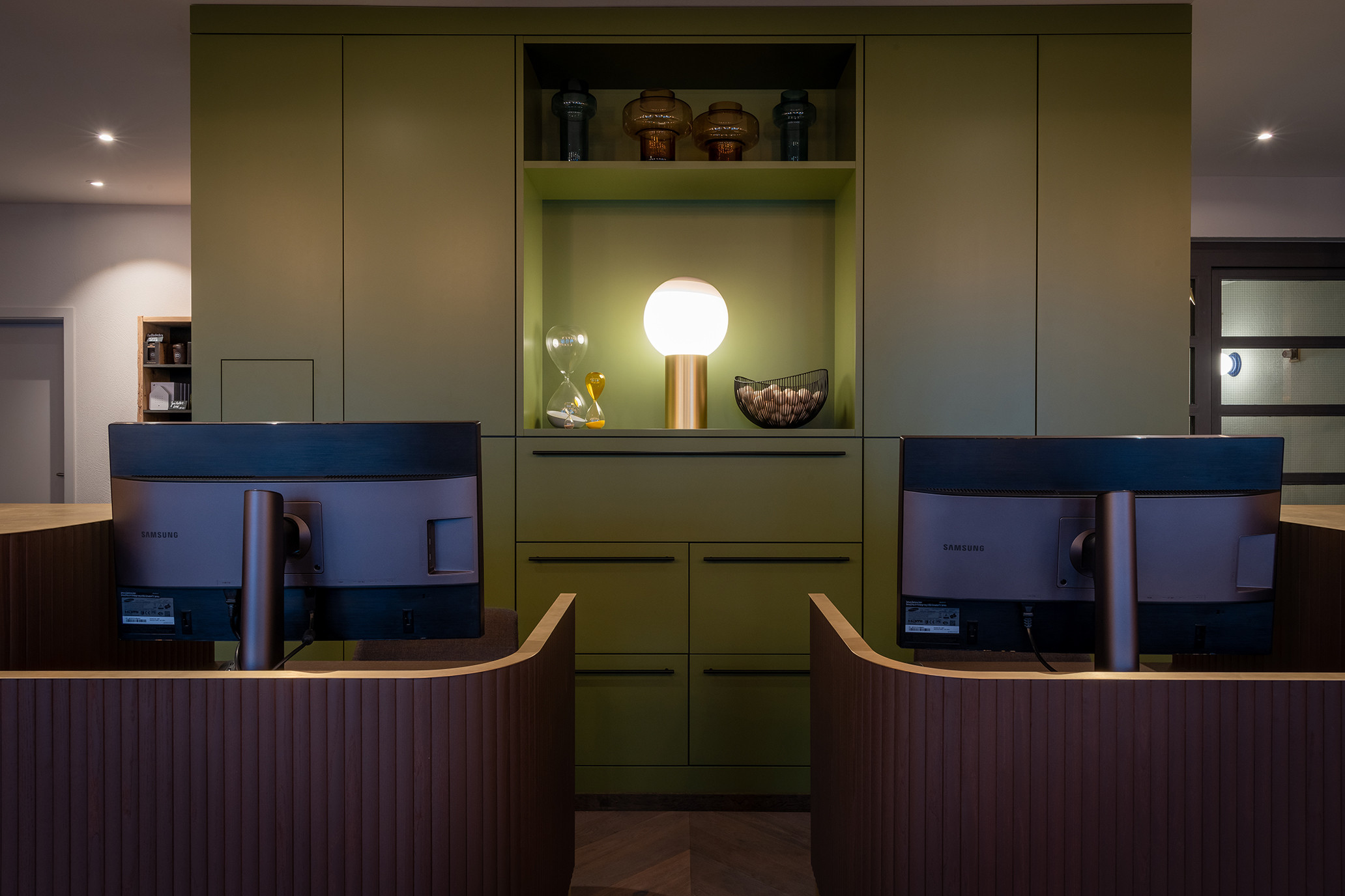 Modern gestalteter Empfangsbereich im Schweizerhof Lenzerheide mit zwei Computerarbeitsplätzen, stilvoller grüner Einbauwand, dekorativer Tischlampe und hochwertigen Design-Accessoires.