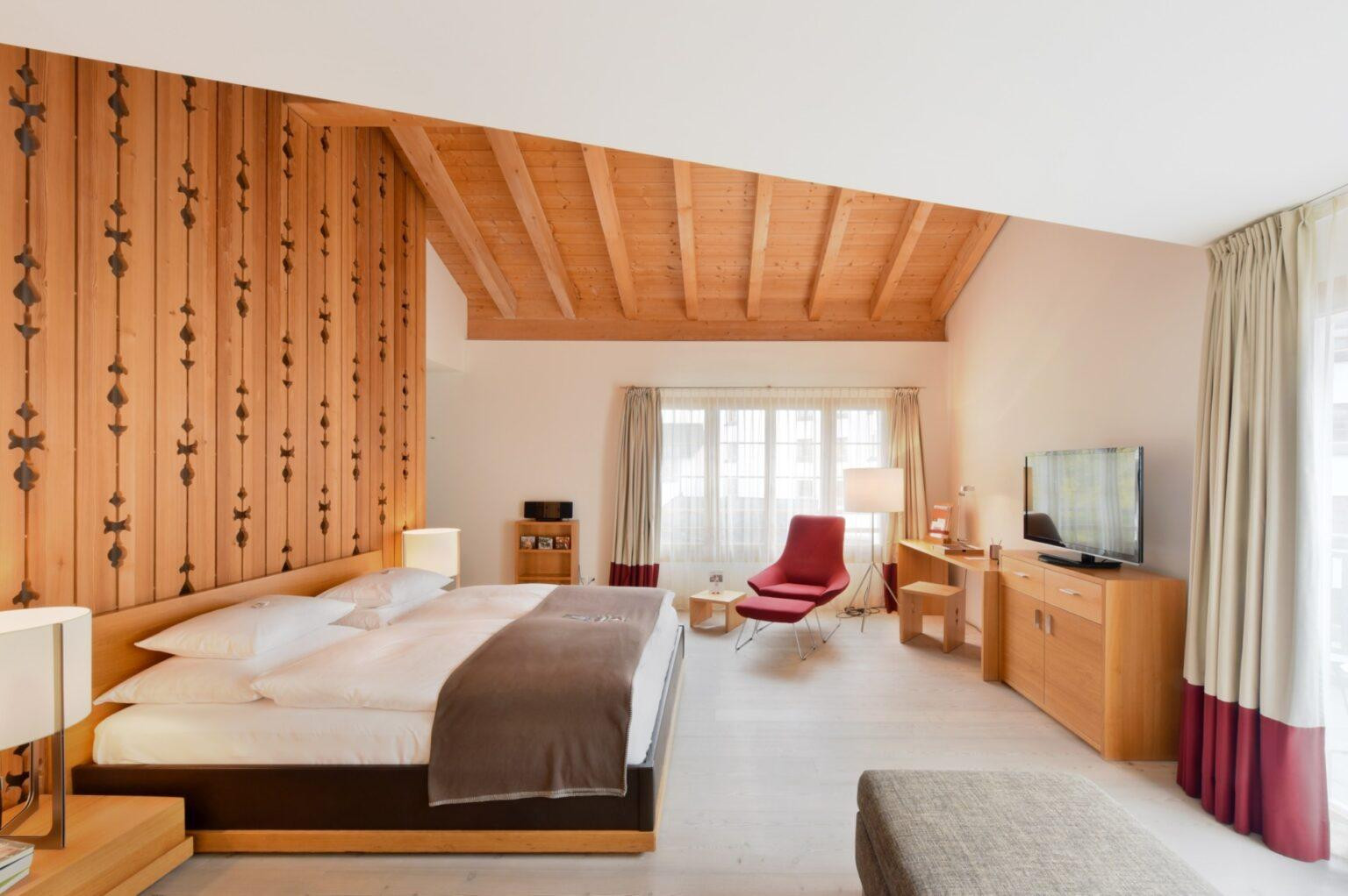 Zweistöckige Alpenchic Suite mit Gallerie, einem Kingsize-Bett, grosse Fenster mit Vorhängen und einer Holzdecke mit Holzbalken und einem Sessel.