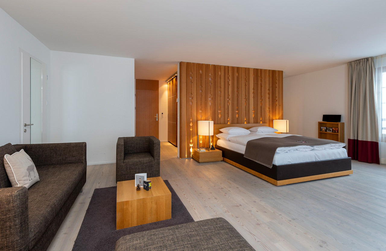 Grosszügige Alpenchic Suite mit Doppelbett vor einer Holzwand, gemütlicher Sitzecke mit Sofas und Beistelltisch.