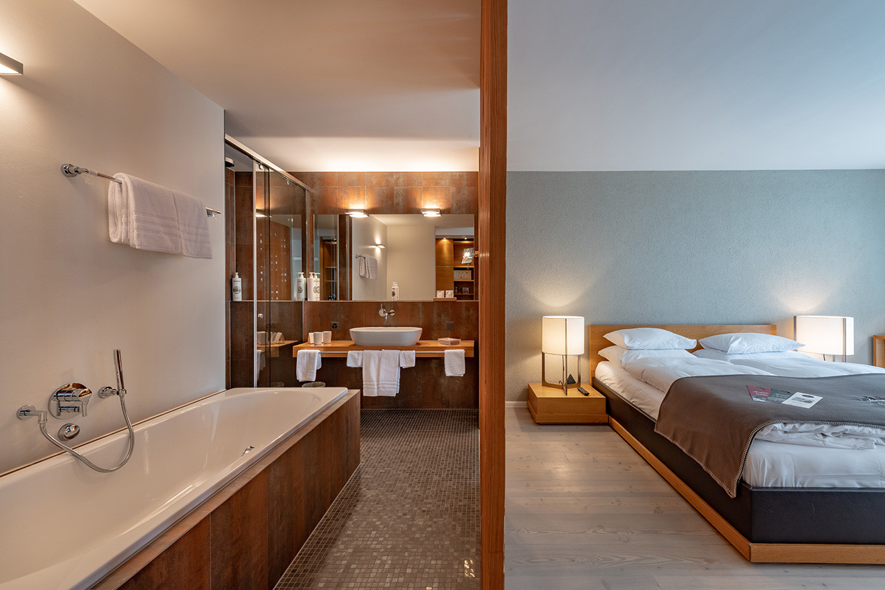 Alpenchic Zimmer welches getrennt wird von der Holztrennwand. Links vom Bild sieht man das Badezimmer mit der Badewanne und das Waschbecken und rechts vom Bild das Bett.