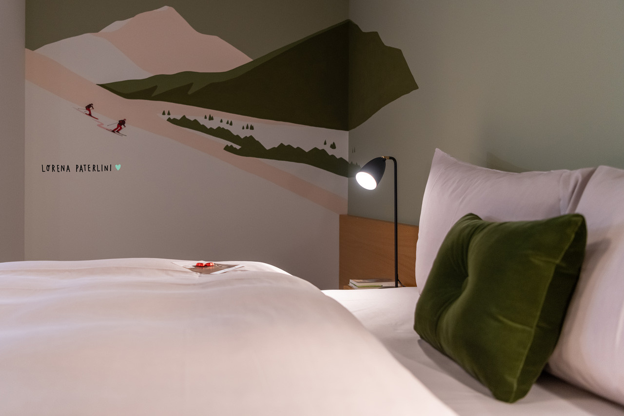 Nahaufnahme seitlich vom Bett mit der Wandmalerei von den Bergen mit Menschen die Ski-fahren von Lorena Paterlini.
